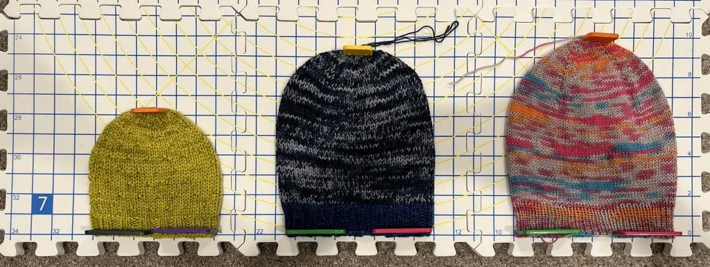Three knitted hats pinned on a gridded blocking mat.
